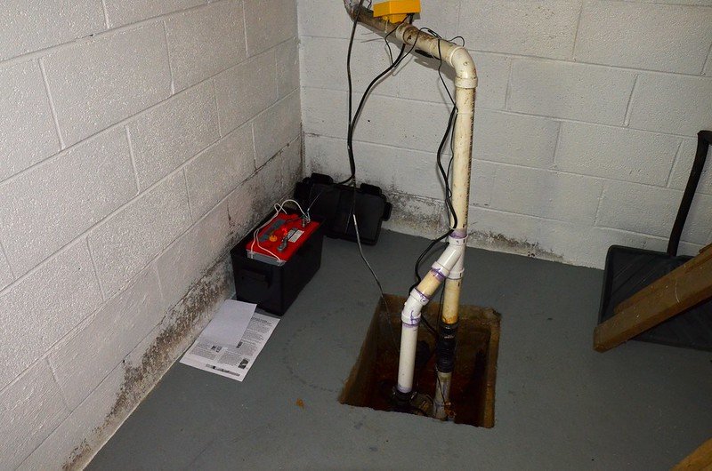 Battery backup sump pump system installed in basement with moisture on walls and exposed wiring – essential for power outage protection